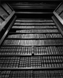 black and white photograph of a tall bookshelf filled with old-fashioned bound book. One book is pulled out a tiny bit.