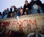 Image of the Berlin Wall with graffiti reading 'FREEDOM' and a group of people standing on top and at the base. A man reaches down to help someone up to join them.