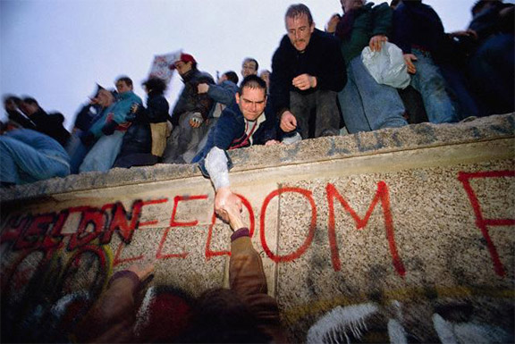 Image of the Berlin Wall with graffiti reading 'FREEDOM' and a group of people standing on top and at the base. A man reaches down to help someone up to join them.