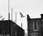Black and white photograph of someone leaping a significant gap between the roofs of two brick buildings.