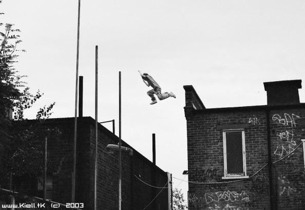 Black and white photograph of someone leaping a significant gap between the roofs of two brick buildings.