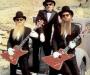 The band ZZ Top standing in a field in front of a beat-up old truck, dressed in tuxedos, sunglasses and white gloves, guitarists holding matching red electric guitars and sporting their trademark long beards.