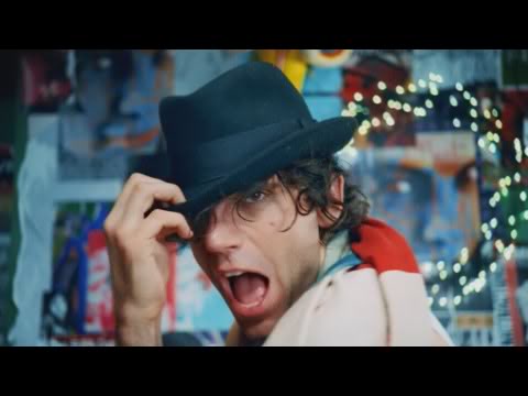 Screencap from Mika's music video of "We Are Golden". Mika is wrapped in a flag, posing with a black hat and singing.