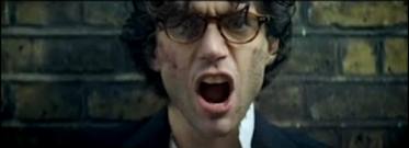 Screen shot from Mika's music video "Kick-Ass". Mika standing defiant against a brick wall in blood and dirt as though he's recently been roughed up.