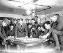 Black and white period photograph of a group of military men crowded around maps on a table. Clearly plotting.
