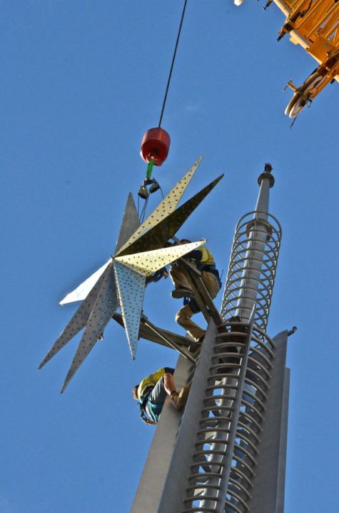 Two people in workman's gear perched on top of the Perth bell tower as a crane lowers the Christmas star into place