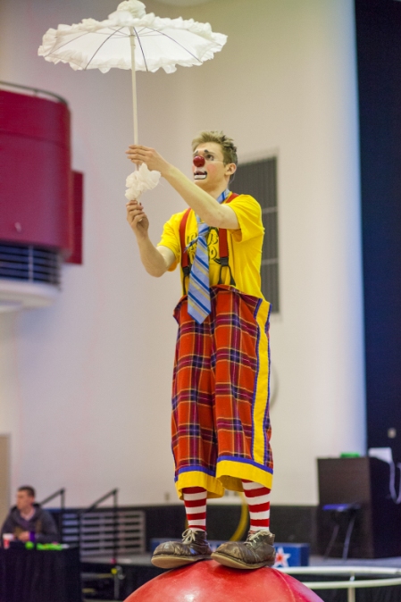 Rather concerned looking clown balancing on a large rubber ball and uncertainly holding a frilly white umbrella.